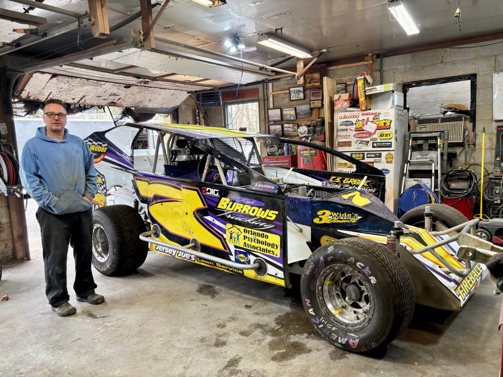 Chris grab standing next to a modified dirt race car in a garage, surrounded by tools, racing memorabilia, and machinery.