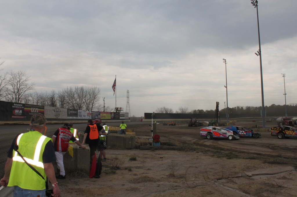 Photographers at work in the middle of the dirt track. They wear bright media jackets and some modified dirt cars may be seen