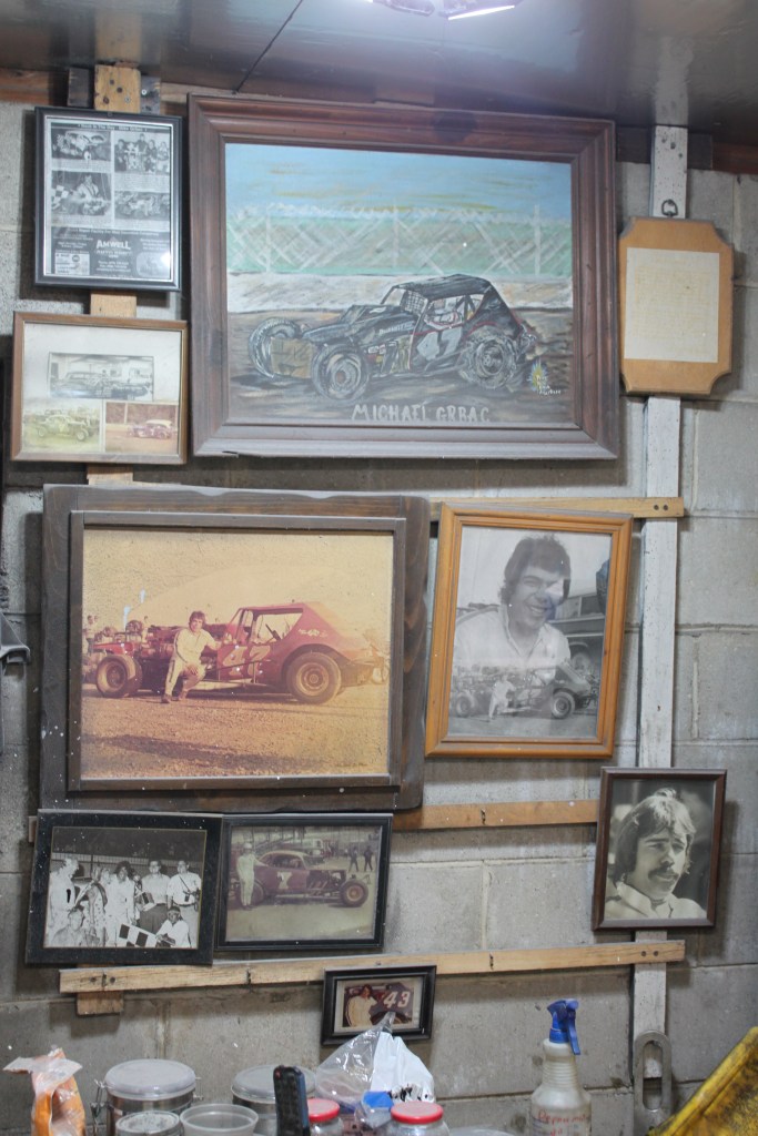 A collection of framed photographs and artwork depicting racing memories, including a painted portrait of Michael Grbac and various vintage racing images on a garage wall.