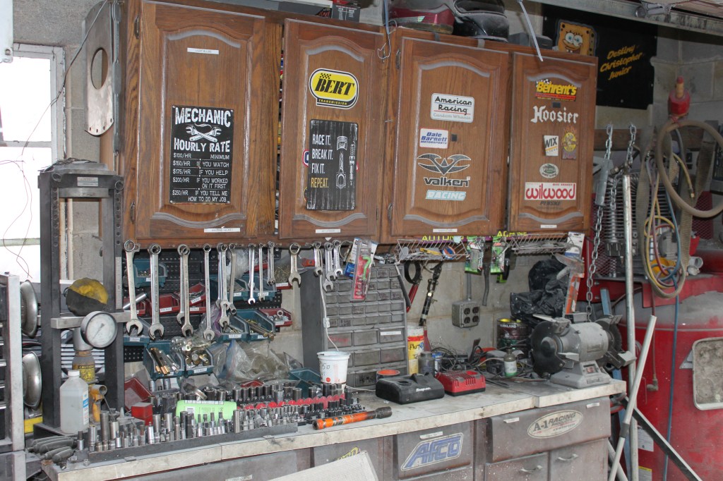 A cluttered garage workspace featuring a workbench filled with various tools, wrenches hanging on the wall, and cabinets adorned with racing stickers, showcasing the environment of a dedicated racer.