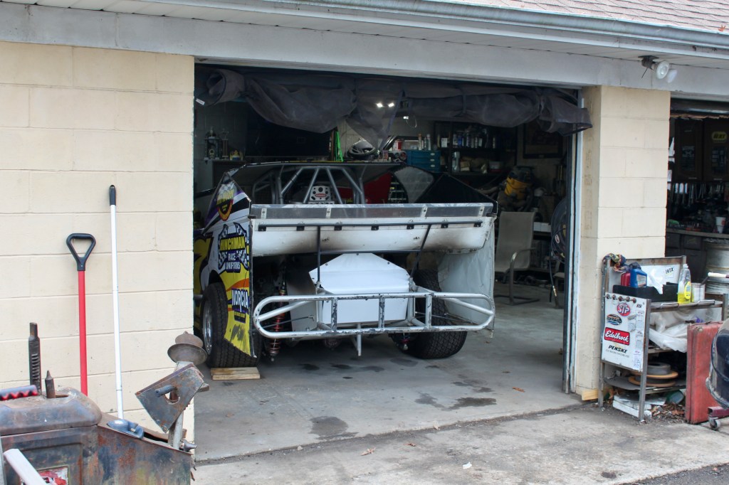 Modified dirt car partially visible in a garage, surrounded by tools and parts, with a shovel leaning against the wall.