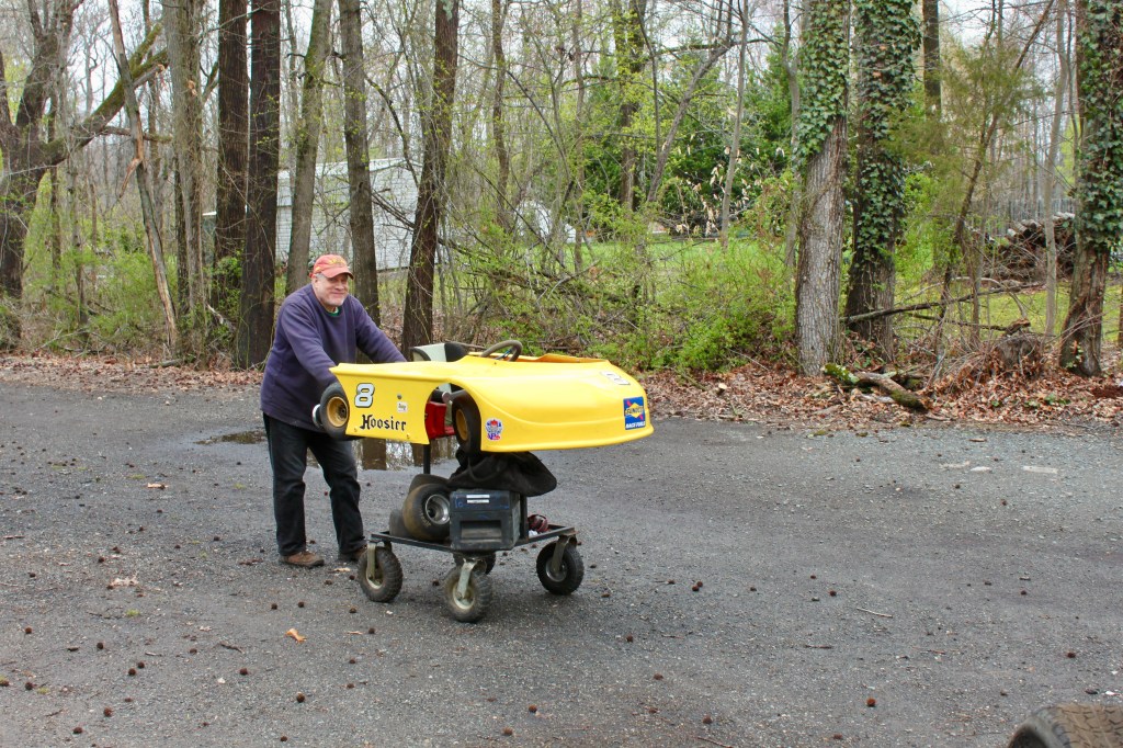 A man pushing a small racing go-kart on a cart in a wooded area, surrounded by trees and fallen leaves.